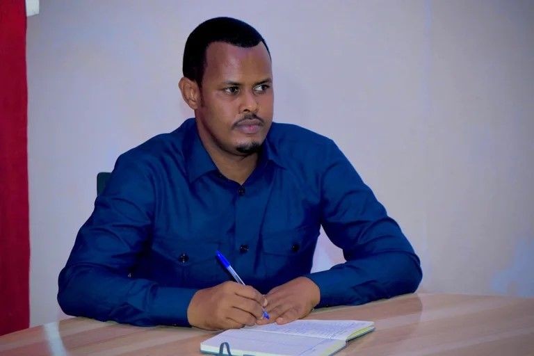 Man in a blue shirt sitting at a desk and writing in a notebook with a blue pen.