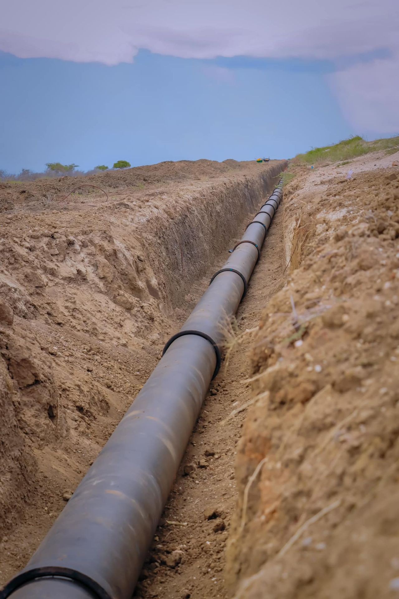 Large black pipeline installed in a deep trench in a dirt field, under a cloudy sky.